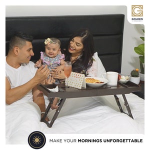 May include: A brown wooden breakfast tray with a white mug, a plate of food, and a glass of pink liquid. The tray is on a white bed with a couple and a baby.