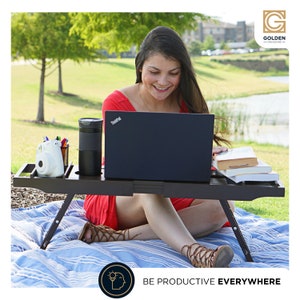 May include: A woman sits on a blanket in a park, working on a laptop on a wooden lap desk. The desk has a built-in cup holder and a small shelf for other items. The woman is wearing a red dress and sandals. The text "BE PRODUCTIVE EVERYWHERE" is visible in the image.