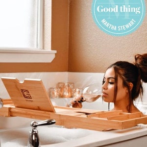May include: A woman relaxes in a bathtub with a bamboo bath tray. The tray has a built-in book holder and a spot for a glass of wine. The tray is approved by Martha Stewart.