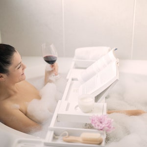 May include: A woman relaxes in a bathtub filled with bubbles, enjoying a glass of red wine and reading a book on a white bath caddy. The caddy has a built-in book holder, a small tray for a candle, and a larger tray for a glass or other items.