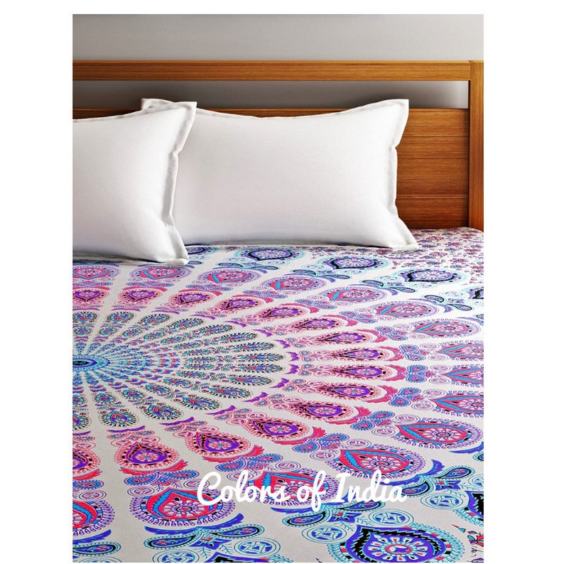 May include: A white bed with two white pillows and a white and blue mandala patterned bedspread. The bedspread has a pink, blue, and purple color gradient. The text "Colors of India" is printed on the bedspread.