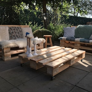 Op de afbeelding: Een houten palletbank met witte en grijze kussens, een bijpassende pallet salontafel met wielen en een kleine houten kruk. De meubels staan op een terras met een groene haag op de achtergrond.