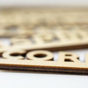 May include: Close-up of a wooden sign with the word "CORN" cut out in the center. The sign is made of light brown wood and has a dark brown edge.