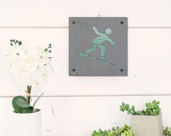 May include: A gray metal sign with a cutout of a person surfing. The person is outlined in blue and green, and the surfboard is outlined in white.