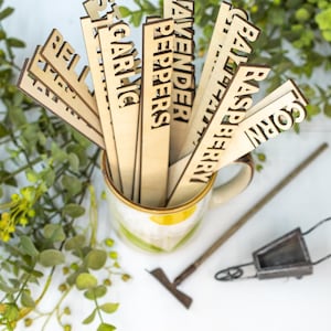 May include: A set of wooden plant markers with various plant names written on them, including "Lavender", "Peppers", "Garlic", "Corn", "Raspberry", and "Bay Leaf". The markers are in a ceramic mug with a floral design.