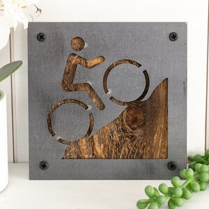 Peut inclure: Une plaque en m&eacute;tal gris avec un motif d&eacute;coup&eacute; d'une personne &agrave; v&eacute;lo montant une colline. La colline est en bois et a une finition brune rustique.
