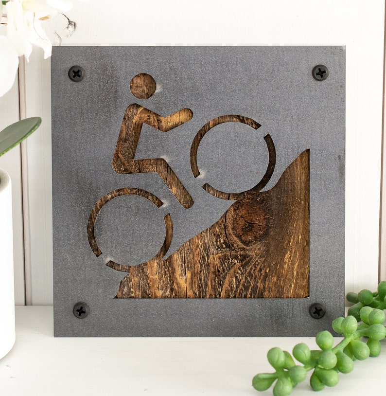 Peut inclure: Une plaque en m&eacute;tal gris avec un motif d&eacute;coup&eacute; d'une personne &agrave; v&eacute;lo montant une colline. La colline est en bois et a une finition brune rustique.