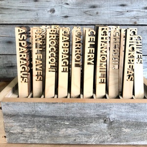 May include: Wooden plant markers with the names of various herbs and vegetables, including asparagus, bay leaves, broccoli, cabbage, carrot, celery, chamomile, chervil, chick peas, chives, corn, and garlic. The markers are arranged in a wooden crate.