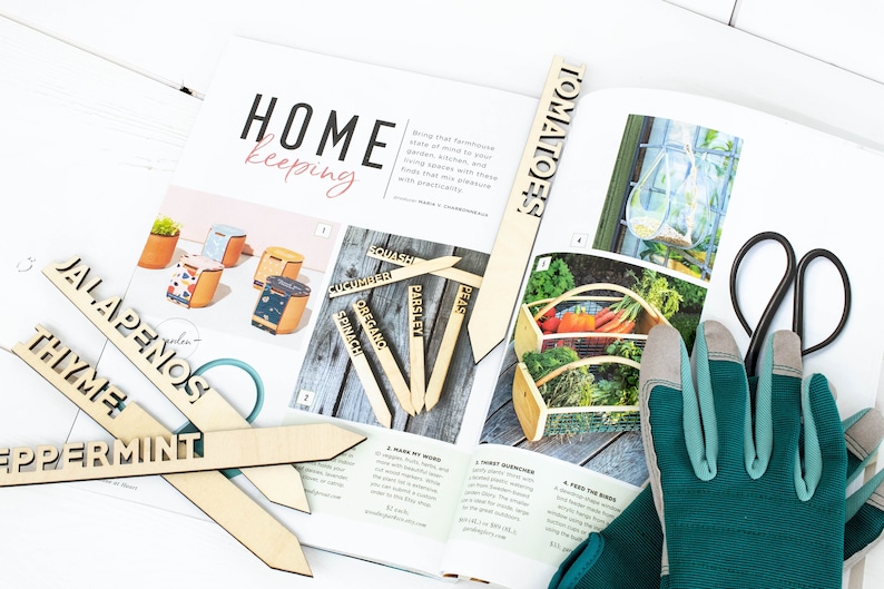 May include: Wooden plant markers with the words "Peppermint", "Thyme", "Jalapenos", "Tomatoes", "Peas", "Parsley", "Spinach", "Oregano", "Cucumber", and "Squash" printed on them. The markers are on a magazine page with a gardening theme.