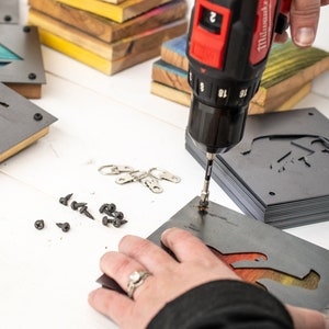 Peut inclure: Une personne utilise une perceuse sans fil rouge et noire pour fixer une plaque de suspension en m&eacute;tal &agrave; une pile de plaques similaires. Les plaques pr&eacute;sentent un motif d&eacute;coup&eacute; d'un buffle. Les plaques sont grises et ont une finition en bois. Il y a aussi des vis et des anneaux de suspension en m&eacute;tal &eacute;parpill&eacute;s sur la table.