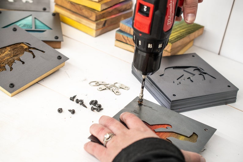 Peut inclure: Une personne utilise une perceuse sans fil rouge et noire pour fixer une plaque de suspension en m&eacute;tal &agrave; une pile de plaques similaires. Les plaques pr&eacute;sentent un motif d&eacute;coup&eacute; d'un buffle. Les plaques sont grises et ont une finition en bois. Il y a aussi des vis et des anneaux de suspension en m&eacute;tal &eacute;parpill&eacute;s sur la table.