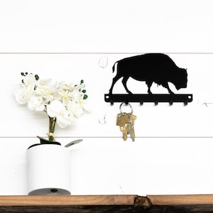May include: Black metal key holder shaped like a buffalo silhouette with five hooks. The key holder is mounted on a white wall with a potted white orchid plant to the left.