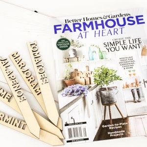 May include: Four wooden plant markers with the words "Tomatoes", "Marjoram", "Jalapenos", and "Mint" written on them. The markers are arranged in a fan shape on a white wooden surface. A magazine titled "Better Homes & Gardens Farmhouse at Heart" is in the background.