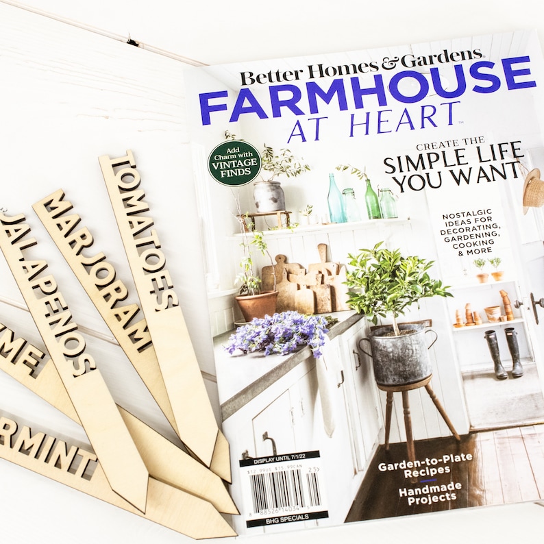 May include: Four wooden plant markers with the words "Tomatoes", "Marjoram", "Jalapenos", and "Mint" written on them. The markers are arranged in a fan shape on a white wooden surface. A magazine titled "Better Homes & Gardens Farmhouse at Heart" is in the background.