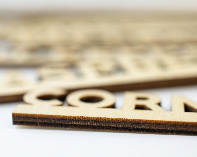 K&ouml;nnte beinhalten: Nahaufnahme eines Holzschildes mit dem ausgeschnittenen Wort "CORN" in der Mitte. Das Schild ist aus hellbraunem Holz und hat einen dunkelbraunen Rand.
