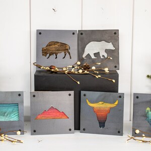Peut inclure: Six d&eacute;corations murales carr&eacute;es en bois avec diff&eacute;rents motifs inspir&eacute;s de la nature. Les motifs comprennent un bison, un ours, une cha&icirc;ne de montagnes, un cr&acirc;ne de taureau et deux contours d'&eacute;tats diff&eacute;rents. Les pi&egrave;ces sont toutes peintes dans un style rustique avec un fond gris et des accents color&eacute;s.