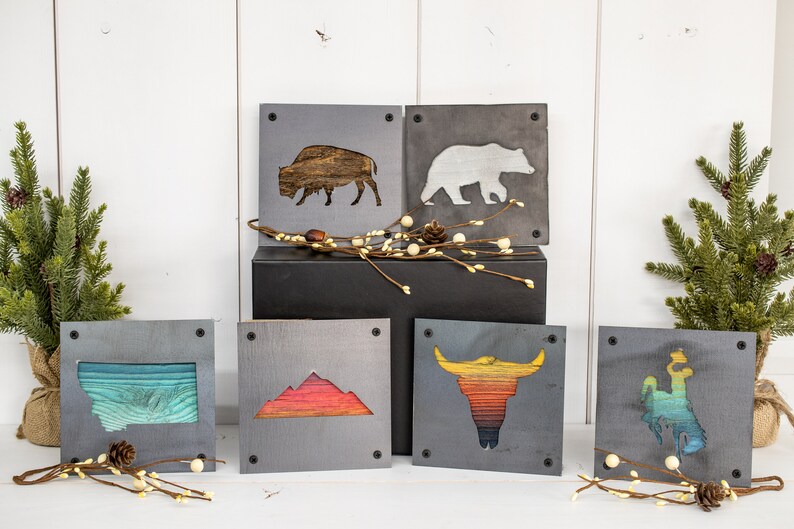 Peut inclure: Six d&eacute;corations murales carr&eacute;es en bois avec diff&eacute;rents motifs inspir&eacute;s de la nature. Les motifs comprennent un bison, un ours, une cha&icirc;ne de montagnes, un cr&acirc;ne de taureau et deux contours d'&eacute;tats diff&eacute;rents. Les pi&egrave;ces sont toutes peintes dans un style rustique avec un fond gris et des accents color&eacute;s.