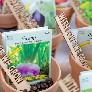 May include: A collection of small terracotta pots with seed packets and wooden signs that say "With Love We Grow". The seed packets are for turnip, cantaloupe, and sweet pea. The turnip seed packet has a picture of a purple top white globe turnip and the text "Heirloom Variety".