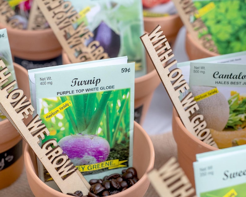 May include: A collection of small terracotta pots with seed packets and wooden signs that say "With Love We Grow". The seed packets are for turnip, cantaloupe, and sweet pea. The turnip seed packet has a picture of a purple top white globe turnip and the text "Heirloom Variety".