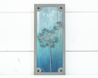 May include: A wood wall art piece with a gray frame and a blue and green gradient background. A metal dandelion design is cut out of the wood.