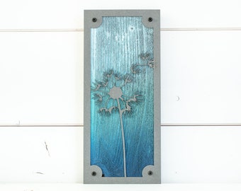 May include: A wood wall art piece with a gray frame and a blue and green gradient background. A metal dandelion design is cut out of the wood.