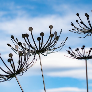 Puede incluir: Tres esculturas metálicas que se asemejan a flores estilizadas con pétalos redondos de color plateado sobre un fondo de cielo azul con nubes blancas.