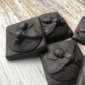 May include: Four black ceramic tiles with raised, detailed images of bees. Each tile features a bee with wings outstretched, set against a textured background.