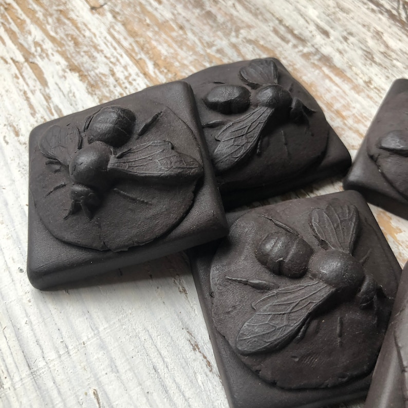 May include: Four black ceramic tiles with raised, detailed images of bees. Each tile features a bee with wings outstretched, set against a textured background.