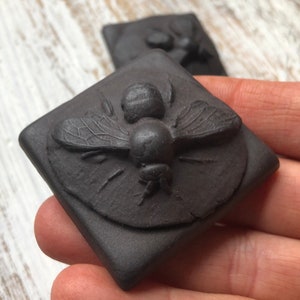May include: A black ceramic tile with a raised design of a bee. The bee is detailed with textured wings and a segmented body.