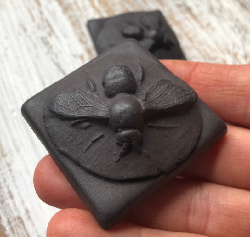 May include: A black ceramic tile with a raised design of a bee. The bee is detailed with textured wings and a segmented body.