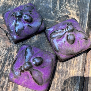 May include: Three purple ceramic tiles with a black bee design. The bees are raised and have a glossy finish.