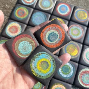 May include: A set of 20 small black square tiles with colorful concentric circles painted on them. The circles are in shades of blue, green, yellow, orange, and red.