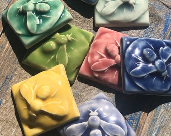 May include: A set of nine ceramic tiles featuring a bee design in various colors, including blue, green, yellow, and pink. Each tile has a raised bee design with intricate details.