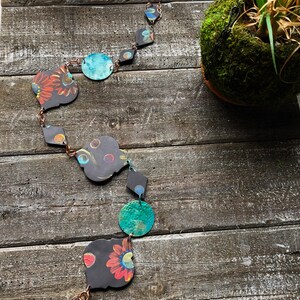 May include: A decorative rain chain featuring dark grey metal shapes in various forms, including circles and diamonds, linked by copper chains. The shapes are adorned with colorful floral patterns. The background is a rustic, weathered wooden surface.