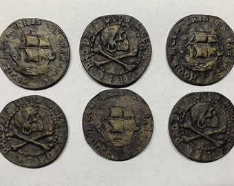 May include: Six antique-looking gold-coloured metal pirate coins with raised images of a skull and crossbones and a ship. The coins have text around the edges.