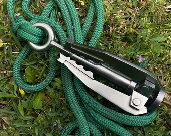 May include: A close-up of a green rope with a metal carabiner clip attached to a black and silver rope tensioner. The tensioner has a serrated edge and a lever.