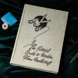Pode incluir: Um livro vintage intitulado "The Pictorial Guide to Modern Home Needlecraft" com uma ilustração a preto e branco de tesouras e linha. Um quadrado azul de sabão, um carretel de linha e pequenas tesouras também estão presentes.