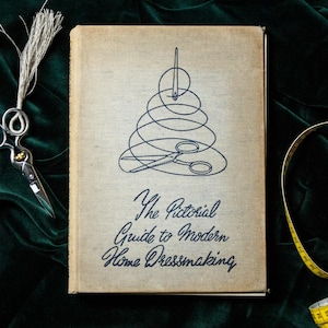 May include: A vintage book titled "The Pictorial Guide to Modern Home Dressmaking" with a needle and thread design on the cover. A pair of silver scissors, a yellow measuring tape, and a green velvet background complete the image.