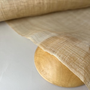May include: A roll of sheer, beige fabric with a fine, open weave. The fabric is draped over a round, wooden object, creating a layered effect. The material has a slightly textured appearance and a subtle sheen.