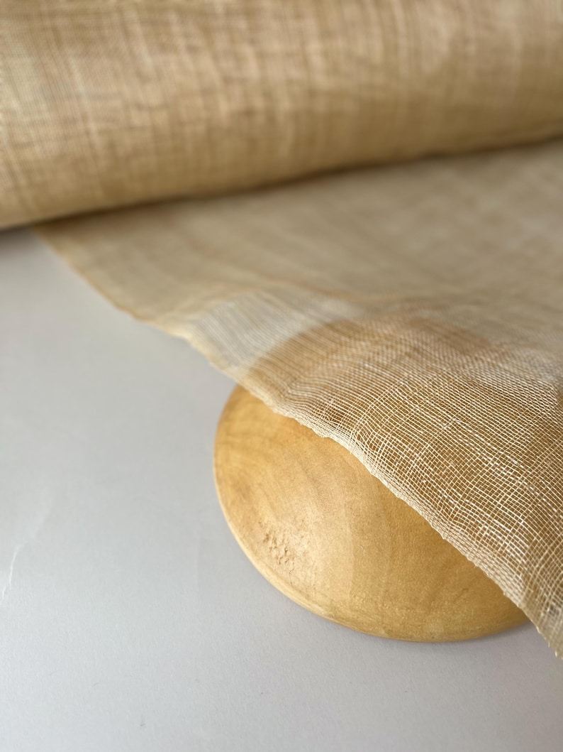 May include: A roll of sheer, beige fabric with a fine, open weave. The fabric is draped over a round, wooden object, creating a layered effect. The material has a slightly textured appearance and a subtle sheen.