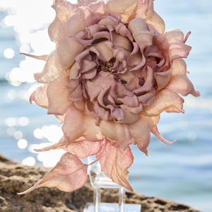 Könnte beinhalten: Eine handgefertigte Stoffblume in Altrosa- und Pfirsichtönen mit geschichteten, ausgefransten Blütenblättern und einer strukturierten Mitte. Die Blume ist auf einem transparenten Acrylständer montiert und zeigt ihre komplexe, organische Form.