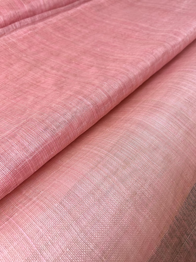 May include: Close-up of folded, light pink fabric with a woven texture. The fabric has a subtle sheen and varying shades of pink, creating depth. The material appears soft and lightweight, suitable for apparel or home decor.