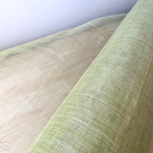 May include: Close-up of two rolls of fabric. One roll is a light green colour, and the other is a beige colour. The fabric has a woven texture, and the rolls are positioned diagonally against a white background.