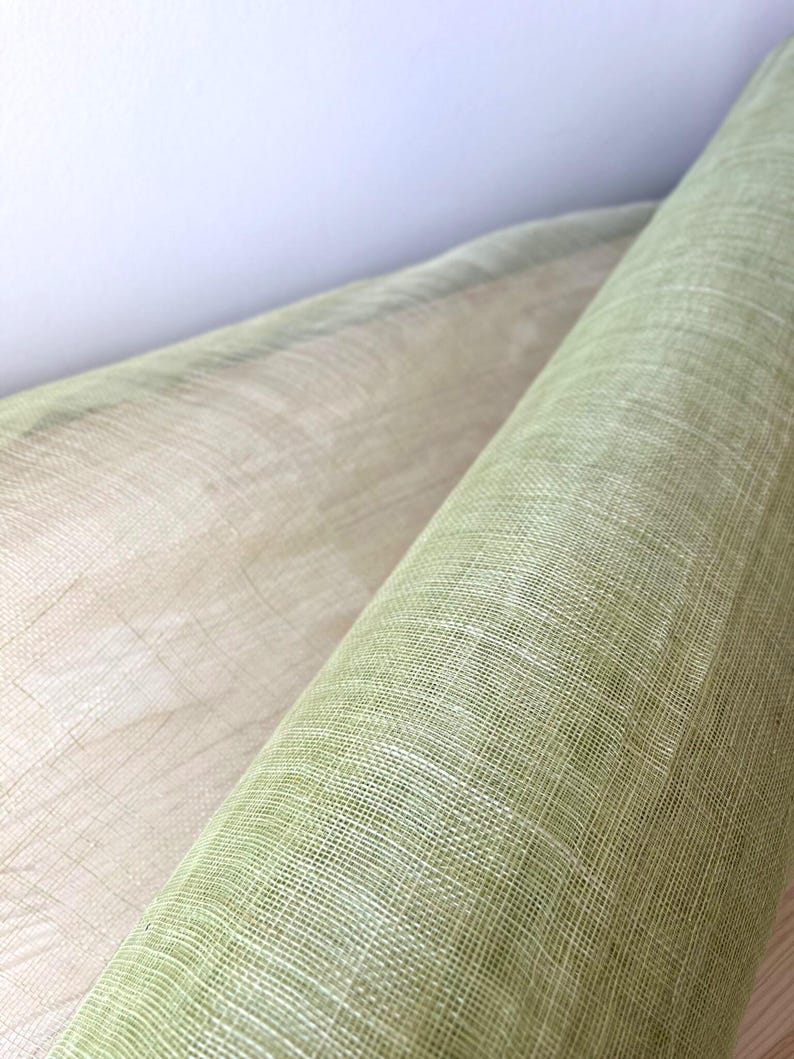 May include: Close-up of two rolls of fabric. One roll is a light green colour, and the other is a beige colour. The fabric has a woven texture, and the rolls are positioned diagonally against a white background.