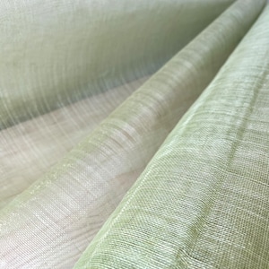 May include: Close-up of a light green fabric with a woven texture. The fabric is folded and draped, revealing layers of varying shades of green and off-white. The material appears to be lightweight and slightly translucent, with a subtle sheen.