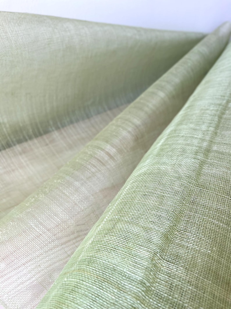 May include: Close-up of a light green fabric with a woven texture. The fabric is folded and draped, revealing layers of varying shades of green and off-white. The material appears to be lightweight and slightly translucent, with a subtle sheen.
