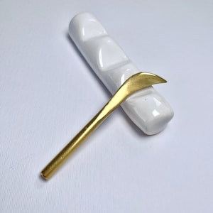 May include: A gold metal spoon with a curved handle rests on a white ceramic dish with three indentations.