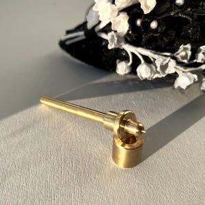 Pode incluir: Uma ferramenta de metal de cor dourada com uma haste longa e fina e uma base redonda e cilíndrica. A ferramenta é projetada para um propósito específico, provavelmente para artesanato ou reparo.