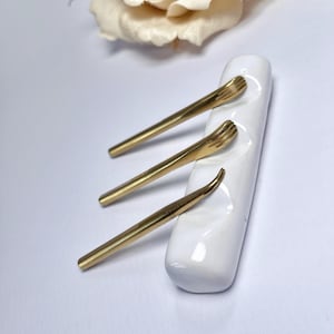 Puede incluir: Tres herramientas de metal dorado descansan sobre un soporte de cerámica blanca. Las herramientas tienen un diseño delgado y alargado con extremos texturizados. Una flor de color crema está en el fondo.