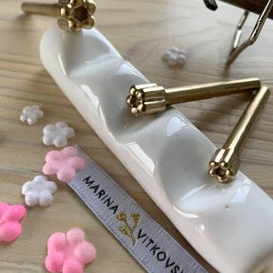 May include: Close-up of decorative hardware. Three gold-colored flower-shaped knobs rest on a white ceramic tray. Small pink and white flower-shaped embellishments are scattered around the tray. A ribbon with the text "MARINA VITKOVSKA" is also present.